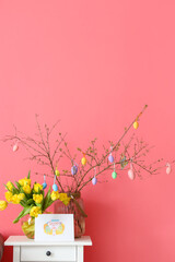 Tree branches with Easter eggs, beautiful tulips and greeting card on table near color wall