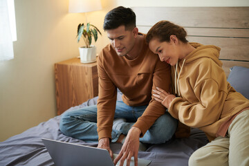 Young couple sitting together on the bed and using laptop to order things online