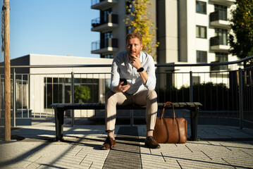 Young stylish businessman sitting on the bench outdoors. Portrait of handsome man using the phone..