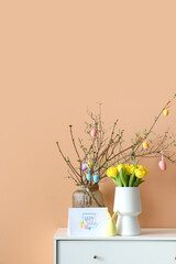Tree branches decorated with painted eggs, tulips and greeting card with text HAPPY EASTER on chest of drawers near color wall