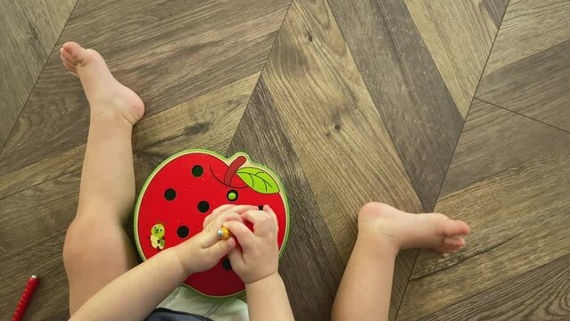 unrecognizable caucasian baby hands playing sorter putting wooden sticks in holes in red apple shape toy. fine motor skills child kid education at home. toddler sitting on brown floor room indoors.