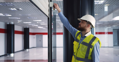 Male chief engineer in hardhat and vest standing inside commercial building construction site, closing an automatic roller door before leaving work.