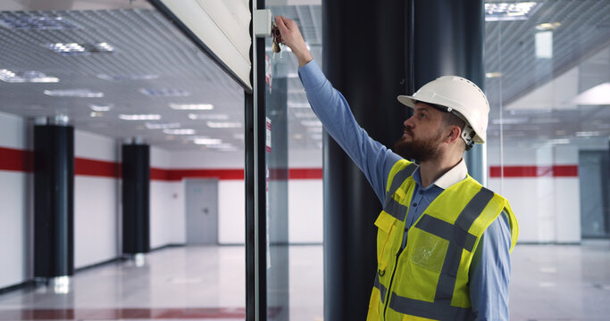 Male chief engineer in hardhat and vest standing inside commercial building construction site, closing an automatic roller door before leaving work.