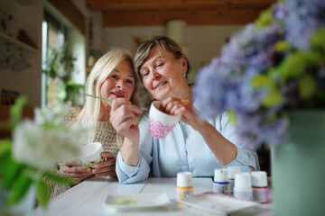 Happy senior woman artist teaching her friend how to paint cermaic cup, artcraft concept.