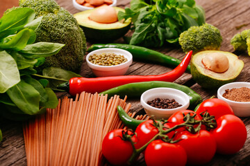 Fresh vegetables for pasta sauce, spaghetti, spices. Green leaves for salad, half of avocado fruit, tomato branch, chilly pepper, organic broccoli. Italian pasta ingredients close up on wooden table