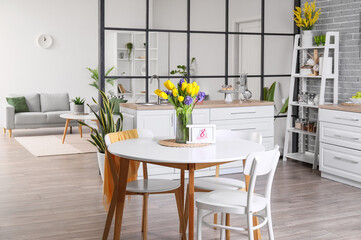 Vase with flowers and greeting card with text 8 MARCH on dining table in kitchen interior. International Women's Day celebration