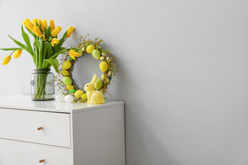 Easter wreath, rabbit and vase with tulips on chest of drawers near grey wall