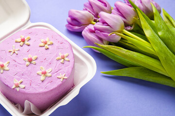 Plastic lunch box with tasty bento cake and flowers on purple background, closeup