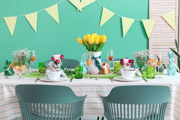Beautiful setting with flowers, burning candles and dinnerware on table served for Easter celebration near color wall
