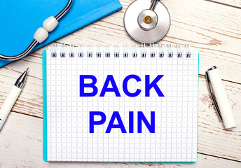 On a light wooden background there is a stethoscope, a blue notebook, a white pen and a sheet of paper with the text BACK PAIN. Medical concept