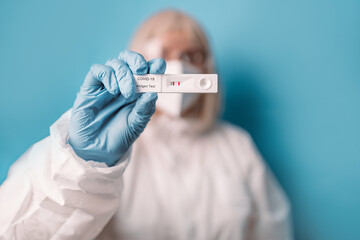 Doctor in medical PPE suit holds Covid-19 rapid antigen test on blue background