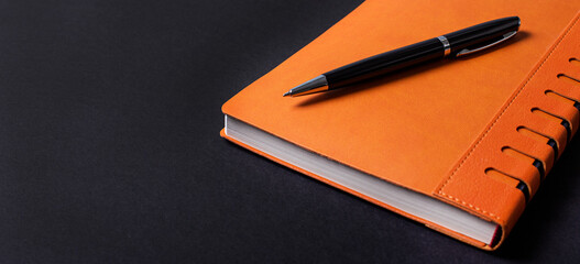 On a black background, an orange notepad and a black pen. Copy space