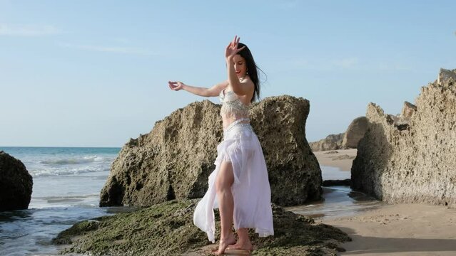 A beautiful woman performing a sensual belly dance in the sand on the beach. Spectacular landscape with blank holes and big rocks. Silhouette.
