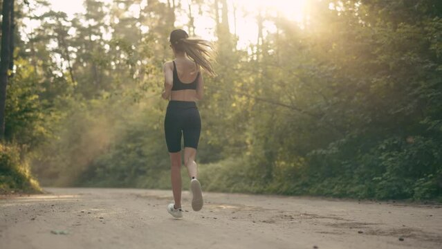 A young woman runner is listening to music in earphones and training in summer forest. The fitness girl is jogging outdoor. Concept of workouts running and healthy lifestyle. Slow motion.