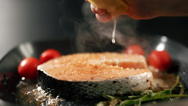 A chef's hand is squeezing a fresh lime over a peace of red sea fish. Salmon fillets with herbs and vegetables is grilling. Cooking process. Concept of healthy food and of freshly made salmon.