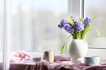 Vase with beautiful hyacinth flowers, cup of tea and candles on windowsill