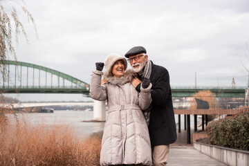 An elderly couple is walking around the waterfront. The couple is smiling and looking happy in love.