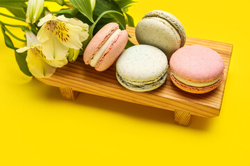 Wooden board with sweet macaroons and flowers on yellow background