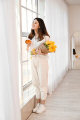 Pretty young Asian woman with bouquet of flowers drinking tea near window