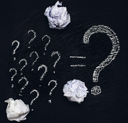 Many small question marks and one large question mark are drawn in white chalk on a rough black...