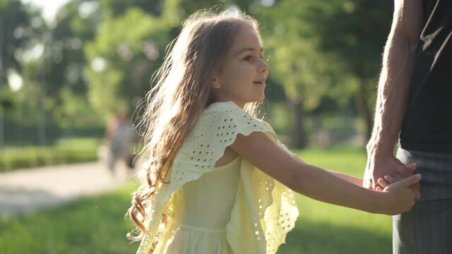 Charming brunette little girl in yellow dress pulling male hand talking standing in sunbeam outdoors. Side view portrait of cute Caucasian daughter resting with unrecognizable father in sunny park