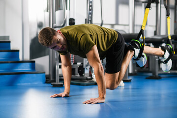 Fototapeta na wymiar Young man making workout exercise for legs with straps at modern gym interior with sports equipment