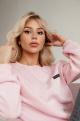 Fashion portrait of beautiful young woman with blonde hair in stylish pastel pastel sweatshirt sits and rest in chair and looks at the camera