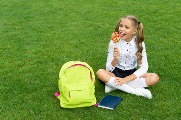 Happy girl child in uniform with school bag and book enjoy eating tasty lollipop sitting on grass