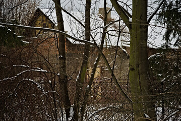 Stary dom z cegły wśród drzew i zarośli , z zasypanym śniegiem dachem kominem , za ogrodzeniem z siatki stalowej.