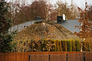 Blaszany dach domu z kominem i antenami , na pierwszym planie domek ( altana , wiata) ogrodowy pokryty strzechą , za płotem ze sztachet i ogrodzeniem z siatki stalowej .