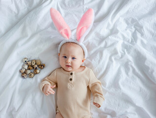 Fototapeta premium Easter costume for children. A cute funny baby with bunny ears costume dressed up for Easter laying on white bed linen sheet with quail eggs. An infant interested looks at the camera.