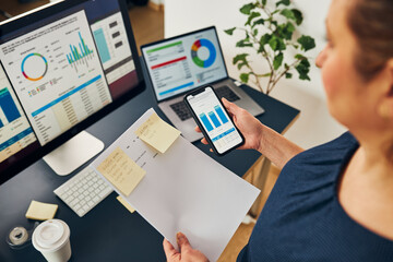 Businesswoman working with financial data on charts and tables on smartphone and computer. Woman entrepreneur looking at sale stats standing by desk in office