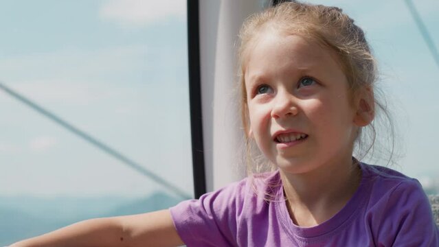 Thoughtful little girl looks up riding gondola with glass windows following moving mechanism on safe poma lift at mountain resort closeup slow motion