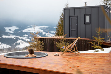 Woman swims in hot bath while resting at small modern house in the mountains on winter. Concept of...