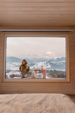 Woman sitting with a drink on terrace of tiny house in the mountains, view through the window from the inside. Concept of small modern cabins for rest and escape to nature