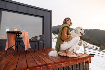 Woman with her dog resting on terrace of tiny house in the mountains on winter time. Concept of small modern cabins for rest and escape to nature. Idea of traveling with dog