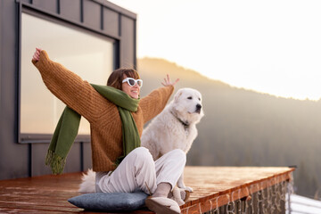 Woman sitting with dog on terrace of tiny house in the mountains enjoying beautiful sunrise landscape. Concept of small modern cabins for rest and escape to nature. Idea of traveling with dog