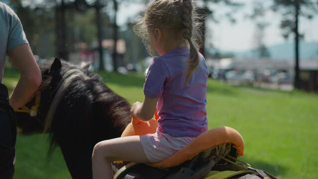 Careful blonde little child girl in lilac t-shirt rides pony with skilled instructor along green city garden on sunny day backside view slow motion