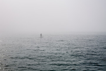 A man on a standing surf rows a paddle far out into the sea in the fog.