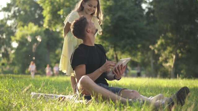 Wide shot relaxed man surfing Internet on tablet as cute little girl covering eyes with hands. Playful Caucasian daughter resting with young handsome father in sunny spring summer park