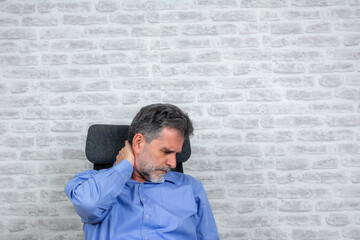unhappy mature man suffering from neck pain at the office, copy space. I need a massage. Frustrated senior man in holding hand on his neck while standing against grey brick wall background
