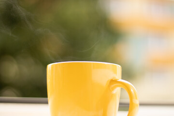 Morning tea on the windowsill from which steam comes out. Yellow cup with a drink near the open window.