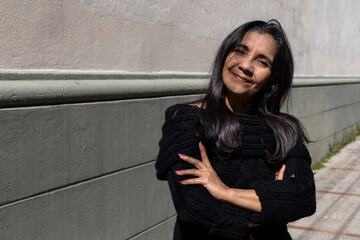 Latin American adult woman (50) poses standing in front of camera with her arms crossed, her...
