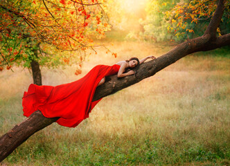 Fantasy woman in long red dress lies on tree branch enjoys nature. Silk fabric of skirt hem flies...