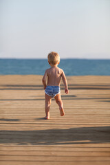 Toddler in striped swimming trunks running along the pier to the sea.Sunset summer time. Beach Vacation holiday concept