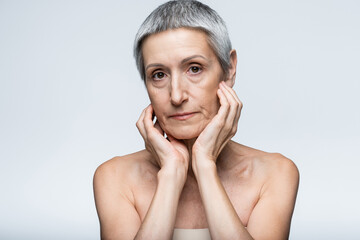 mature woman with bare shoulders touching cheeks and looking at camera isolated on grey