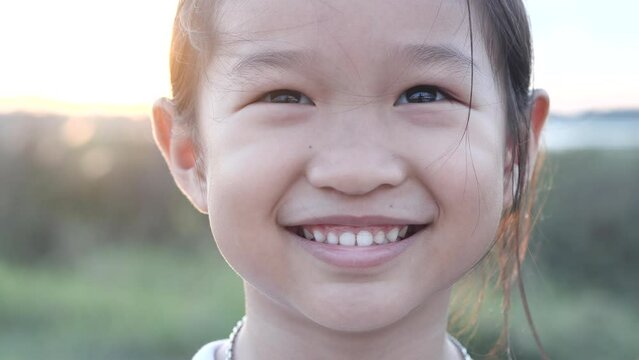 Portrait happy Asian little child girl smiling, laughing, looking at camera with beautiful sunset or sunrise in summer or spring nature outdoor background. Concept of portrait, happy children, family.