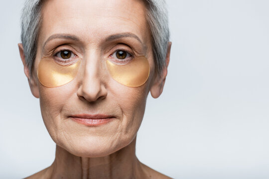 close up of middle aged woman in collagen eye patches looking at camera isolated on grey
