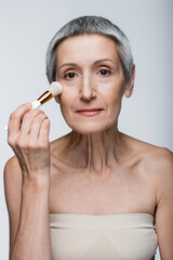 mature woman with grey hair applying face powder with cosmetic brush isolated on grey