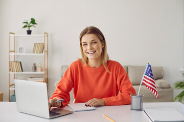 Portrait of happy young woman with USA flag learning American English online from home using laptop. Smiling female student sitting at table and taking online educational course in foreign languages.